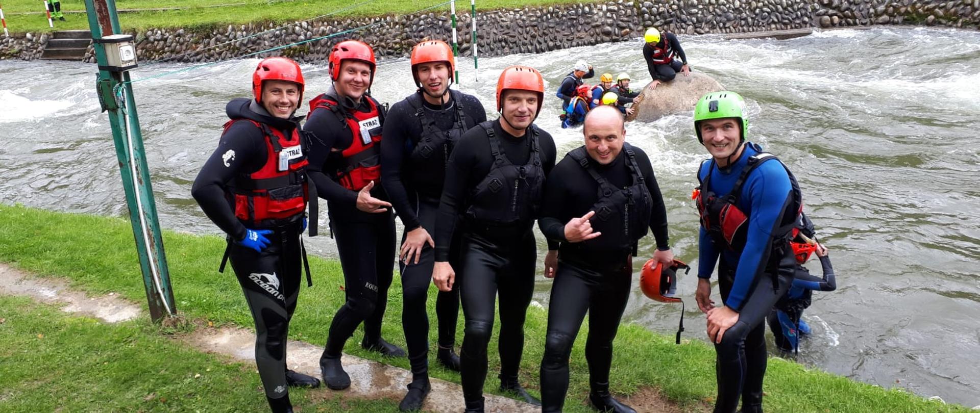 Na zdjęciu znajdują się strażacy ubrani w kamizelki asekuracyjne oraz pianki, w tle widać tor wodny oraz innych strażaków