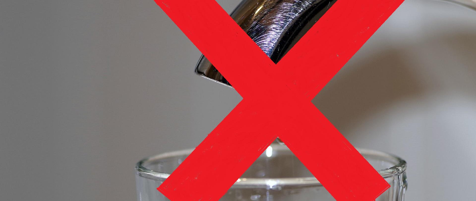 Szklanka wypełniona wodą stojąca pod kranem przekreślona czerwonym krzyżem symbolizująca zakaz picia wody z kranu 