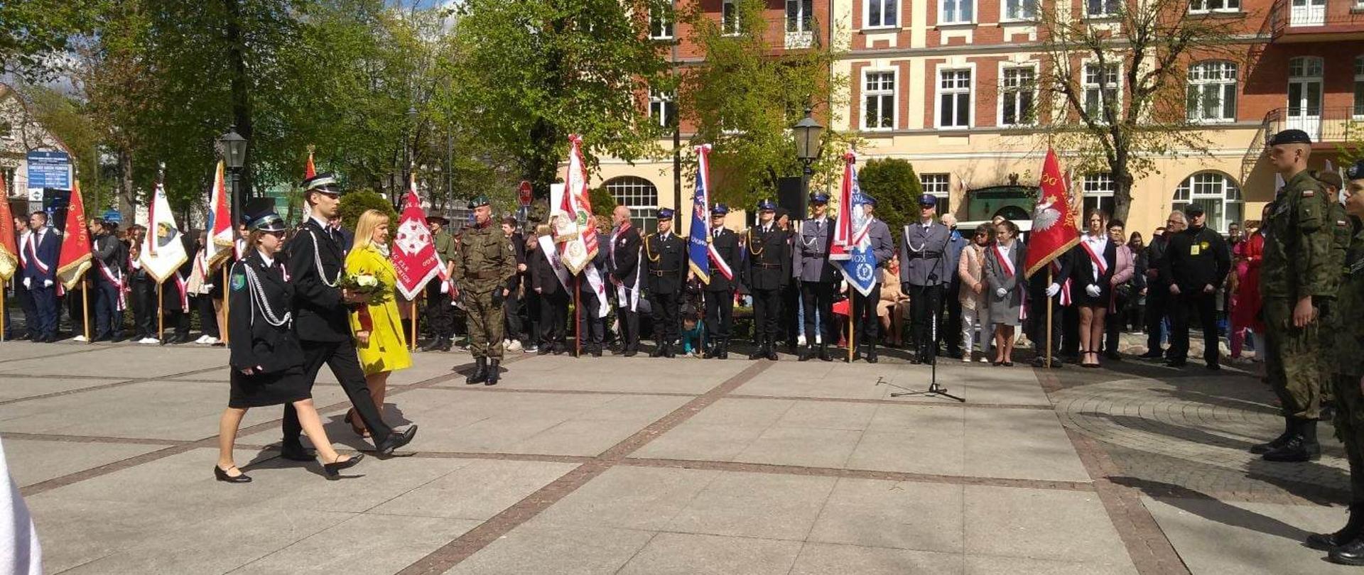 Zdjęcie przedstawia dziewczynę i chłopaka w mundurach Ochotniczej Straży Pożarnej oraz nauczycielkę w żółtym płaszczu. Trójka osób składa kwiaty pod pomnikiem poświęconym Poległym za Wolną i Niezawisłą Polskę. W tle delegacje grup ze sztandarami.