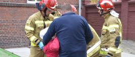 Wypadek podczas prac polowych. Teren jednego z zakładów pracy na terenie Rawicza. Strażacy udzielają kwalifikowanej pierwszej pomocy kobiecie z urazem ręki. Poszkodowaną przytrzymuje mężczyzna w ubraniu cywilnym. W tle budynek i mur z czerwonej cegły.