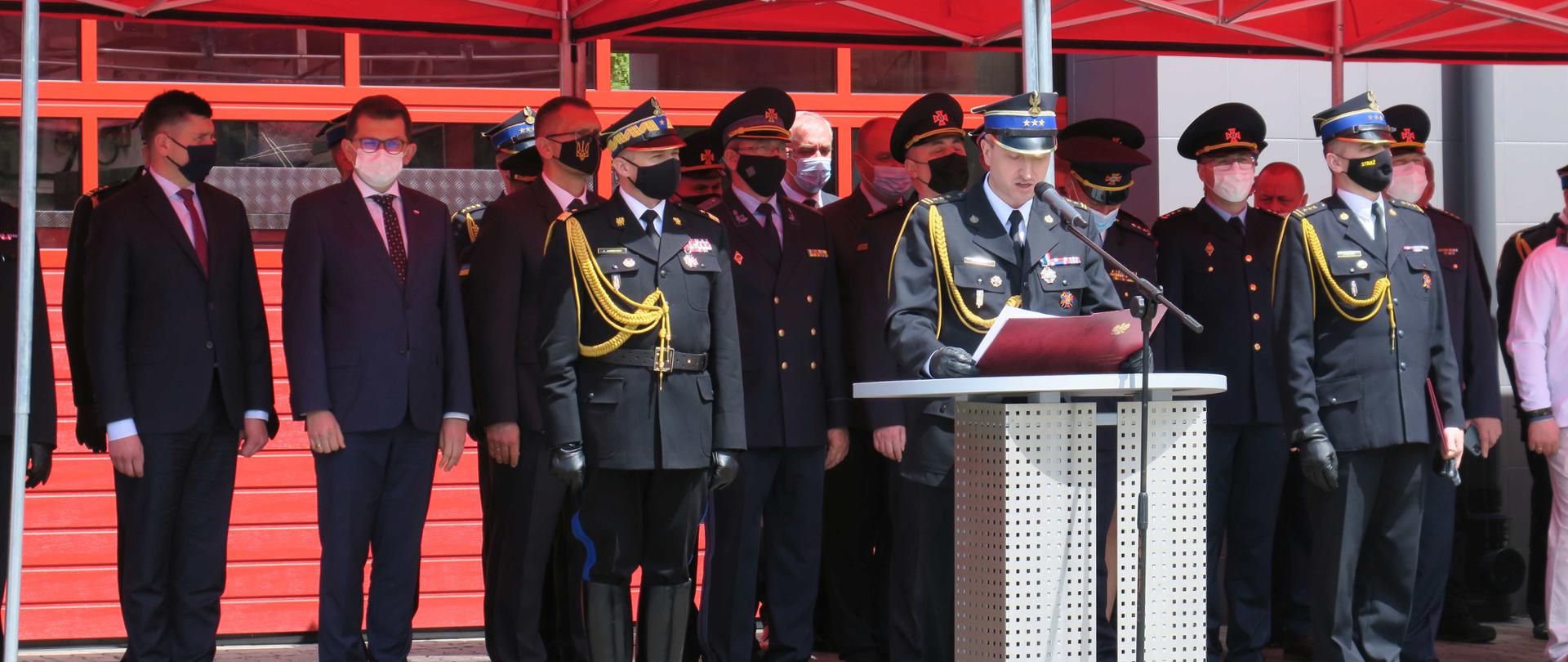 Trybuna honorowa przed której pod czerwonym baldachimem stoją strażacy w mundurach galowych. Przemawia małopolski komendant wojewódzki PSP.