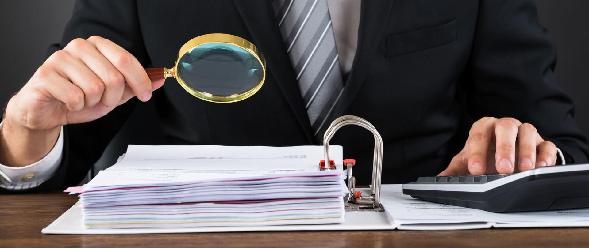 Dłoń trzyma lupę nad segregatorem z dokumentami, druga dłoń obsługuje kalkulator.