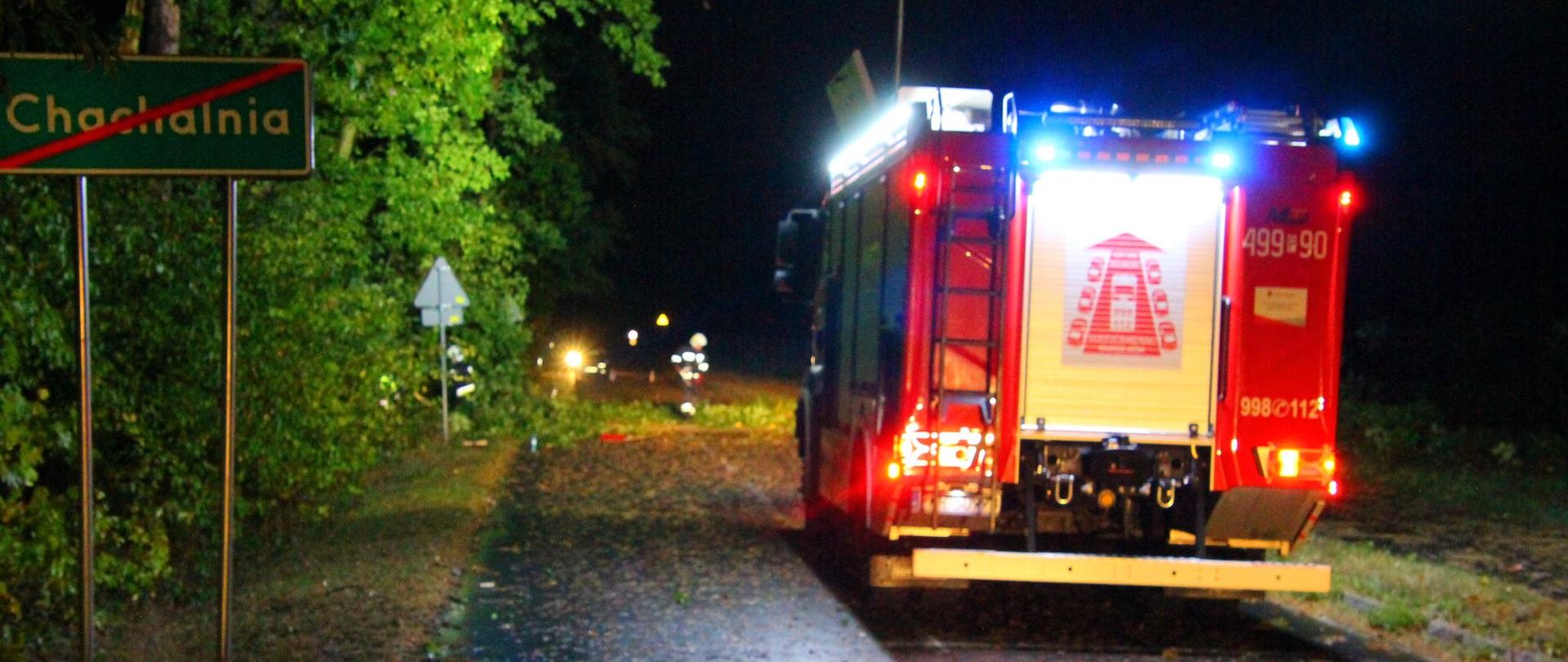 Zdjęcie przedstawia samochód pożarniczy stojący na drodze, na poboczu znak z przekreśloną nazwą miejscowości CHACHALNIA, strażacy piłami mechanicznymi tną leżące na drodze drzewo.