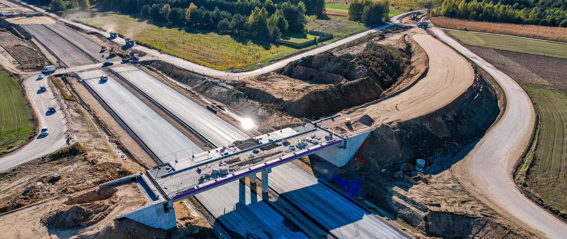 Widok z lotu ptaka na budowę drogi ekspresowej S19 w rejonie Malewice–Chlebczyn. Na zdjęciu widać szerokie pasy nowej jezdni w trakcie układania nawierzchni, most nad trasą z zamontowanymi barierami oraz otaczające nasypy ziemne i drogi techniczne. W tle lasy i pola.