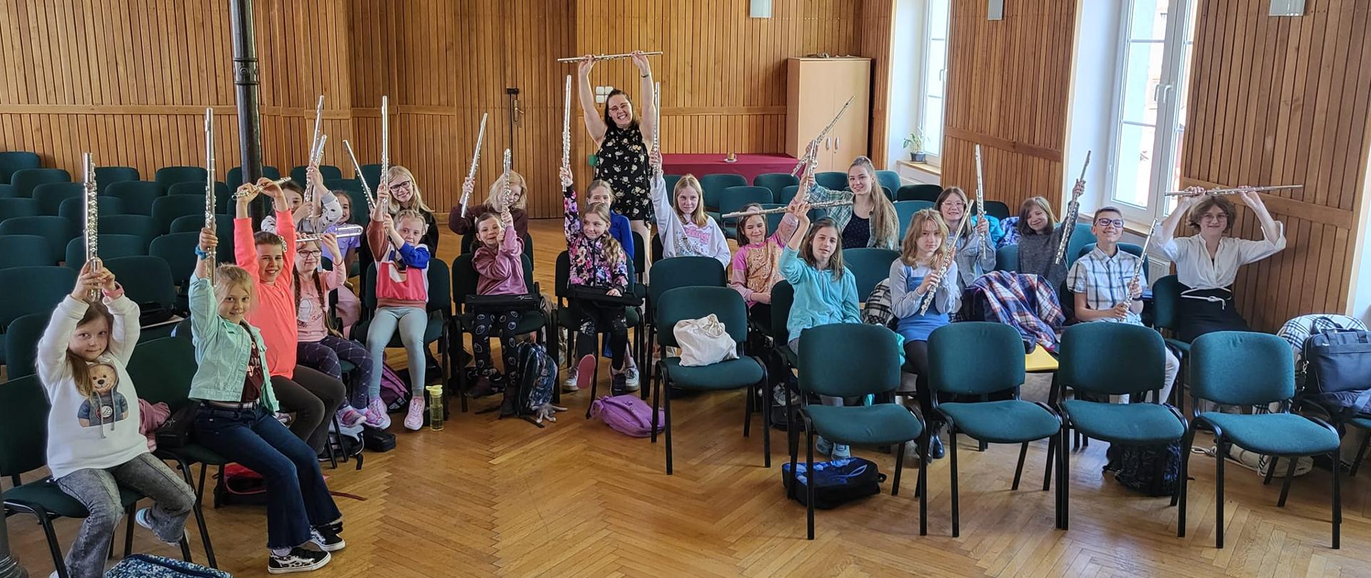 Fotografia przedstawia nauczyciela wraz z uczniami na sali koncertowej, podczas warsztatów fletowych
