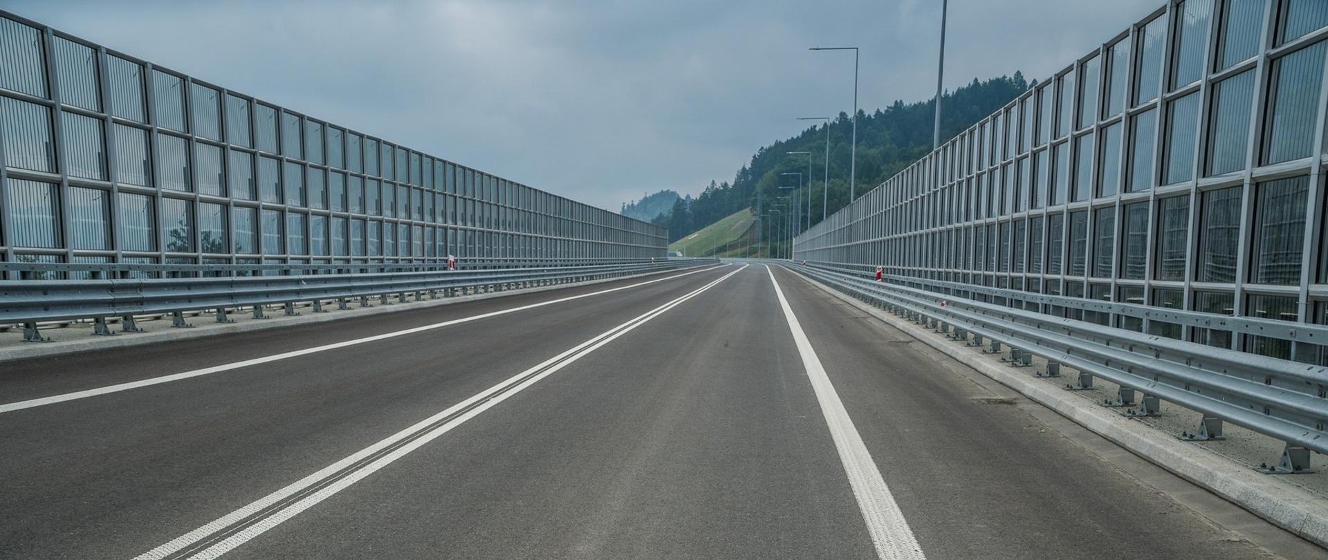 Droga rozpościera się wśród górskich okoliczności natury. Na fotografii dostrzec możemy nawierzchnię drogi jednojezdniowej, z oznakowaniem poziomym oraz ekranami akustycznymi przeziernymi po lewej i prawej stronie. Drogę oświetlać będą latarnie w technologii LED.