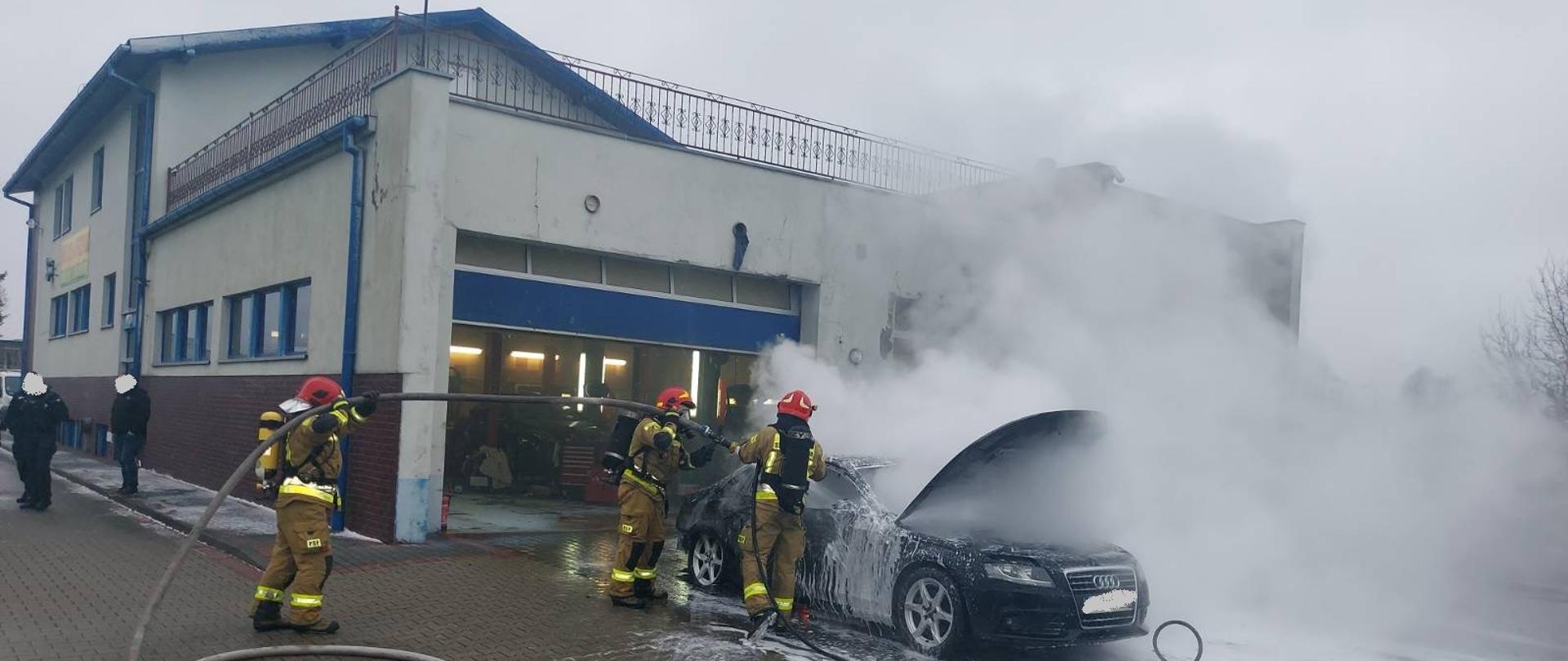Zdjęcie przedstawia palący się samochód osobowy, który zaparkowany jest przed budynkiem warsztatu samochodowego. Strażacy gaszą pożar podając wodę oraz pianę na palący się pojazd. Panuje zadymienie. Strażacy pracują w sprzęcie ochrony układu oddechowego. 