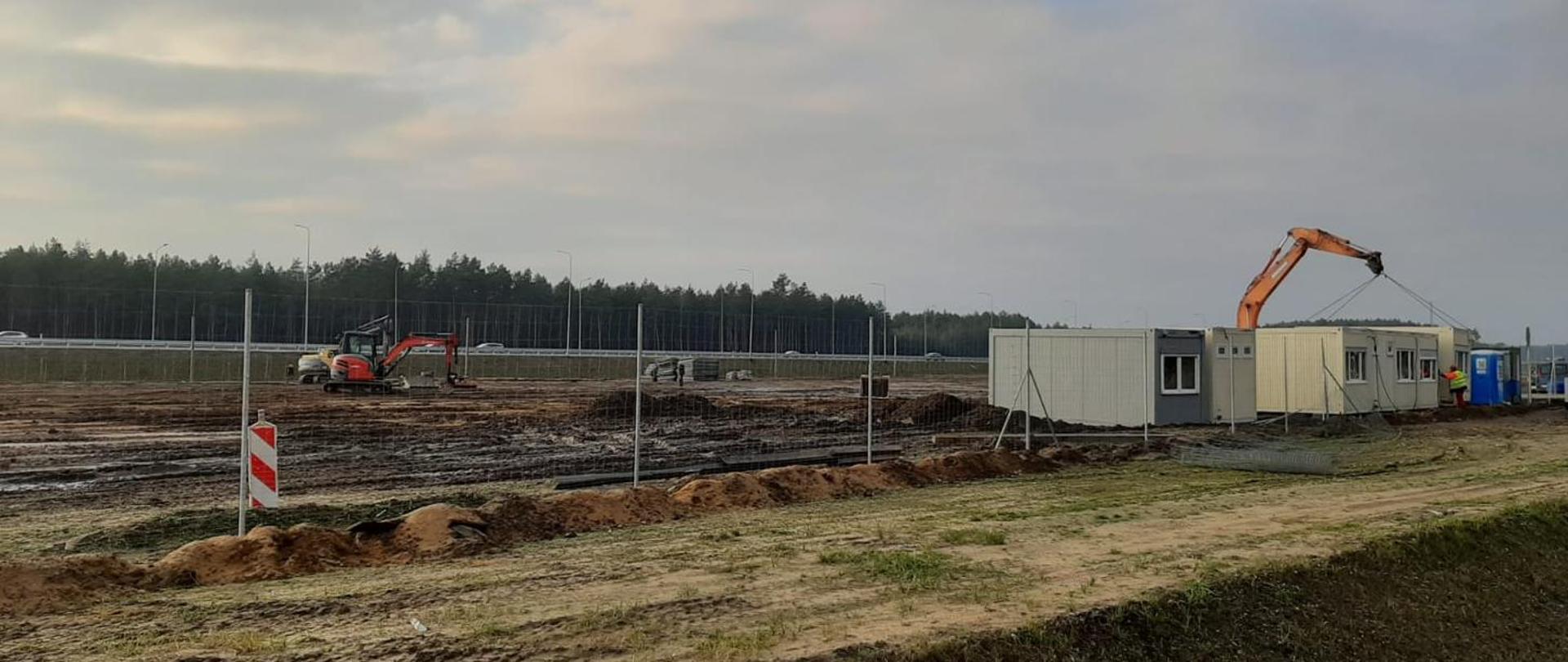 Trwające roboty budowlane. Ogrodzony plac pod przyszłe MOP na S3. Pracujący sprzęt budowlany. Widoczne kontenery mieszkalne robotników budowlanych.