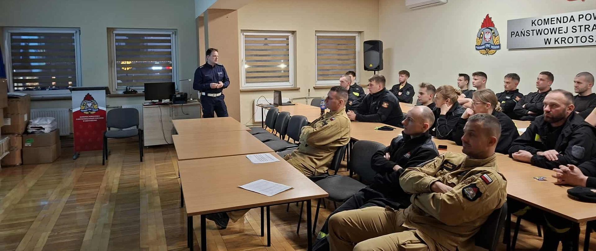 Funkcjinariusz Policji prowadzi wykład dla uczestników szkolenia kierowców konserwatorów z zakresu bezpieczeństwa ruchu drogowego. Za pomocą prezentacji multimedialnej omawia zagadnienia przydatne dla strażaków kierowców pojazdów uprzywilejowanych. Spotkanie ma miejsce na świetlicy komendy. Straacy zasiadają w ławkach w wsłuchują się w zagadnienia poruszane przez Policjanta.