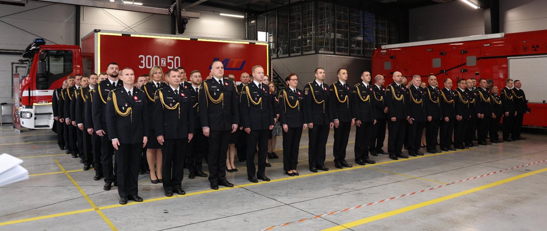 Na zdjęciu widać dużą grupę strażaków, którzy stoją prosto. Wszyscy mają ciemne mundury.
Strażacy mają też na ramieniu złote sznury. Uroczystość odbywa się w dużym, wysokim budynku. W tle, za strażakami, widać dwa samochody strażackie.
