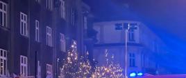 Zdjęcie wykonane w porze nocnej na miejskiej ulicy. Na ulicy samochody służb ratowniczych. Posiadają sygnalizację ostrzegawczą. W tle kamienica. W kadrze ujęta również latarnia oświetlenia ulicznego.