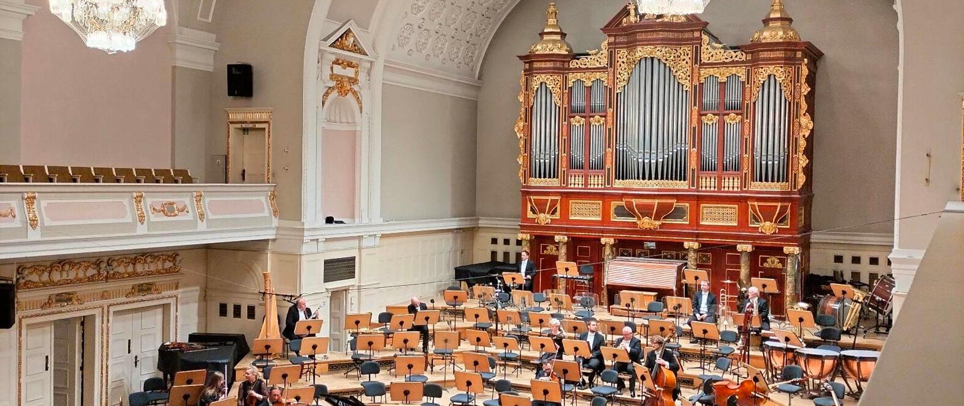zdjęcie wnętrza filharmonii, po prawej organy, u dołu muzycy na scenie, pulpity, krzesła oraz widownia , po lewej balkon, u góry dwa żyrandole