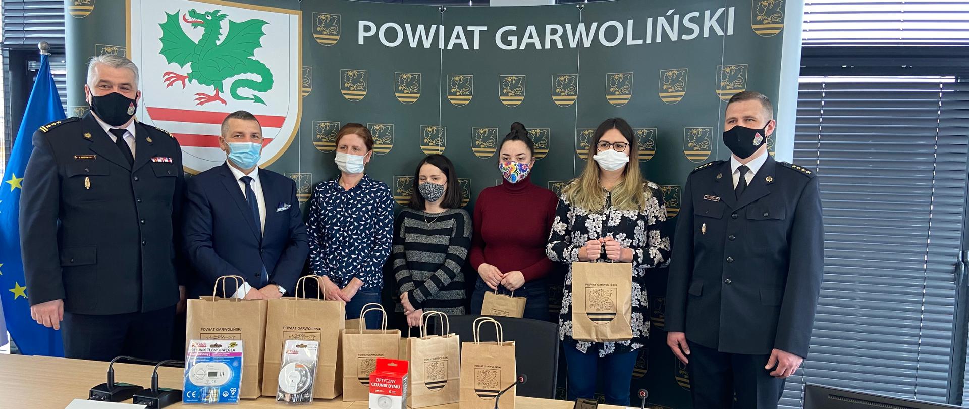 Grupa osób stojąca na tle baneru z logiem powiatu garwolińskiego, Przed osobami na stole ustawione torby z czujnikami tlenku węgla.