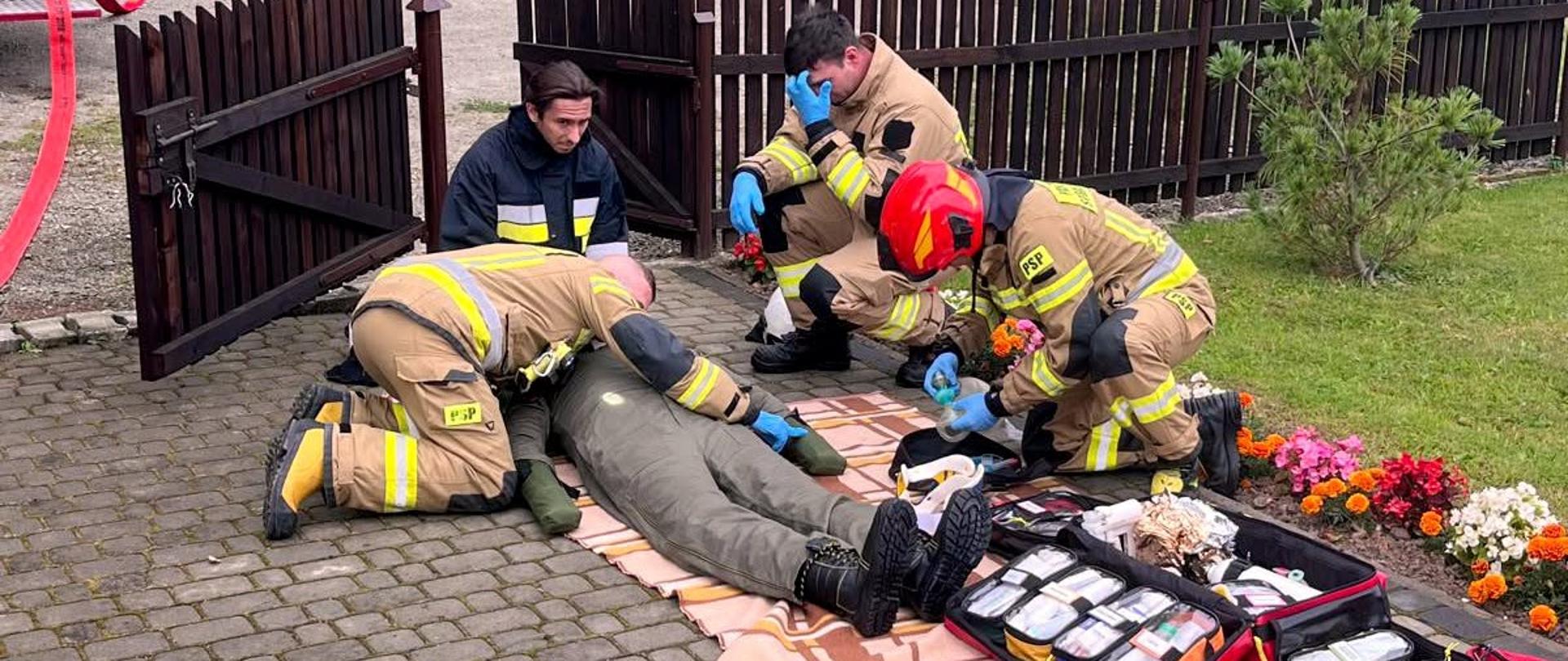 Zdjęcie przedstawia plac przed kościołem. Przed ogrodzeniem stoją dwa samochody pożarnicze. Przy ogrodzeniu strażacy udzielają kpp na fantomie. 