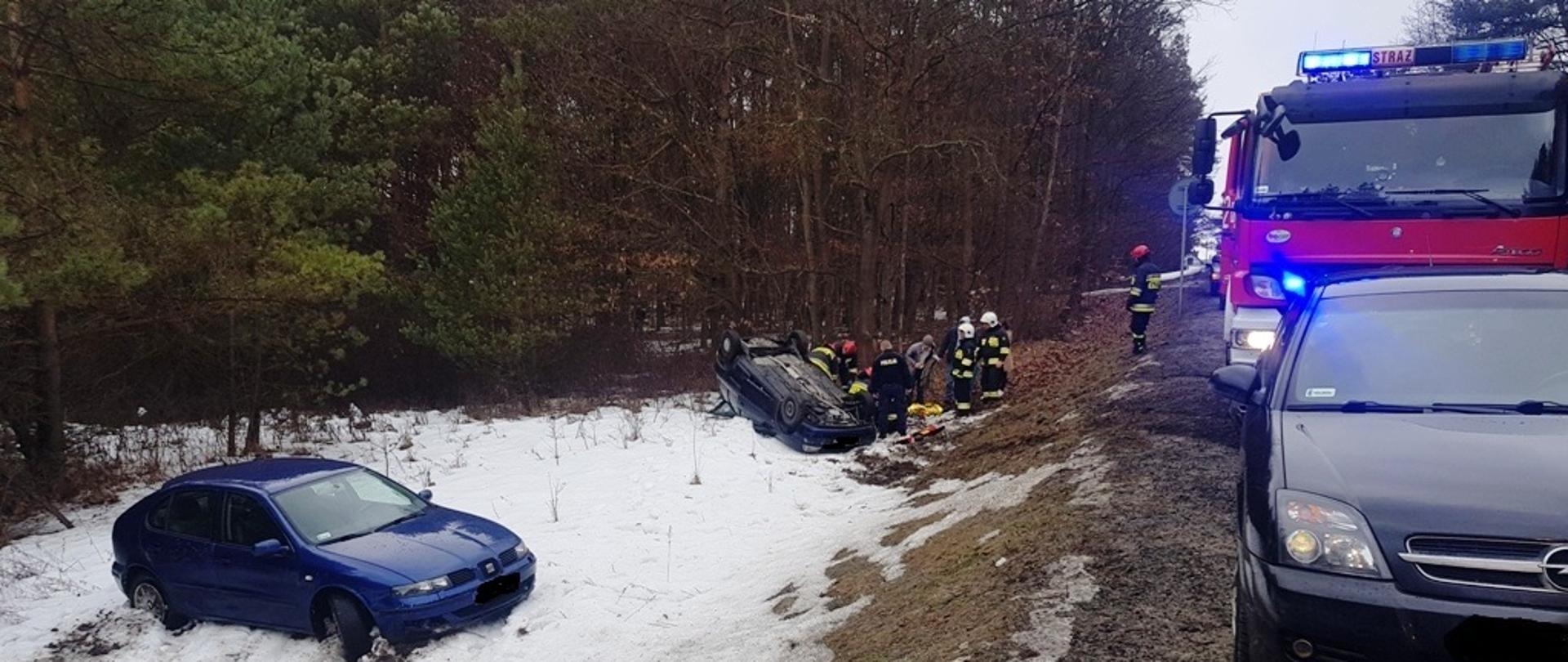 Na zdjęciu widać pojazdy w rowie przy drodze, ratowników udzielających pomocy oraz samochód staży pożarnej jak również samochód osobowy stojące na drodze w pobliżu krańca jezdni