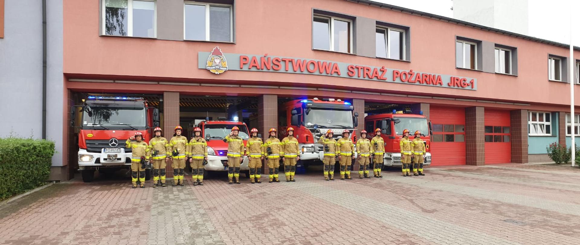 Na zdjęciu widać funkcjonariuszy PSP w Kaliszu, którzy stoją w szeregu przed garażami JRG 1 na ulicy Nowy Świat. Funkcjonariusze mają piaskowy kolor mundurów i czerwone hełmy. Za plecami ratowników w szeregu stoją wyprowadzone z garażu samochody strażackie. Samochody są w kolorze czerwonym. Nad autami znajduje się napis „Państwowa Straż Pożarna – JRG1”. Po lewej logo Państwowej Straży Pożarnej