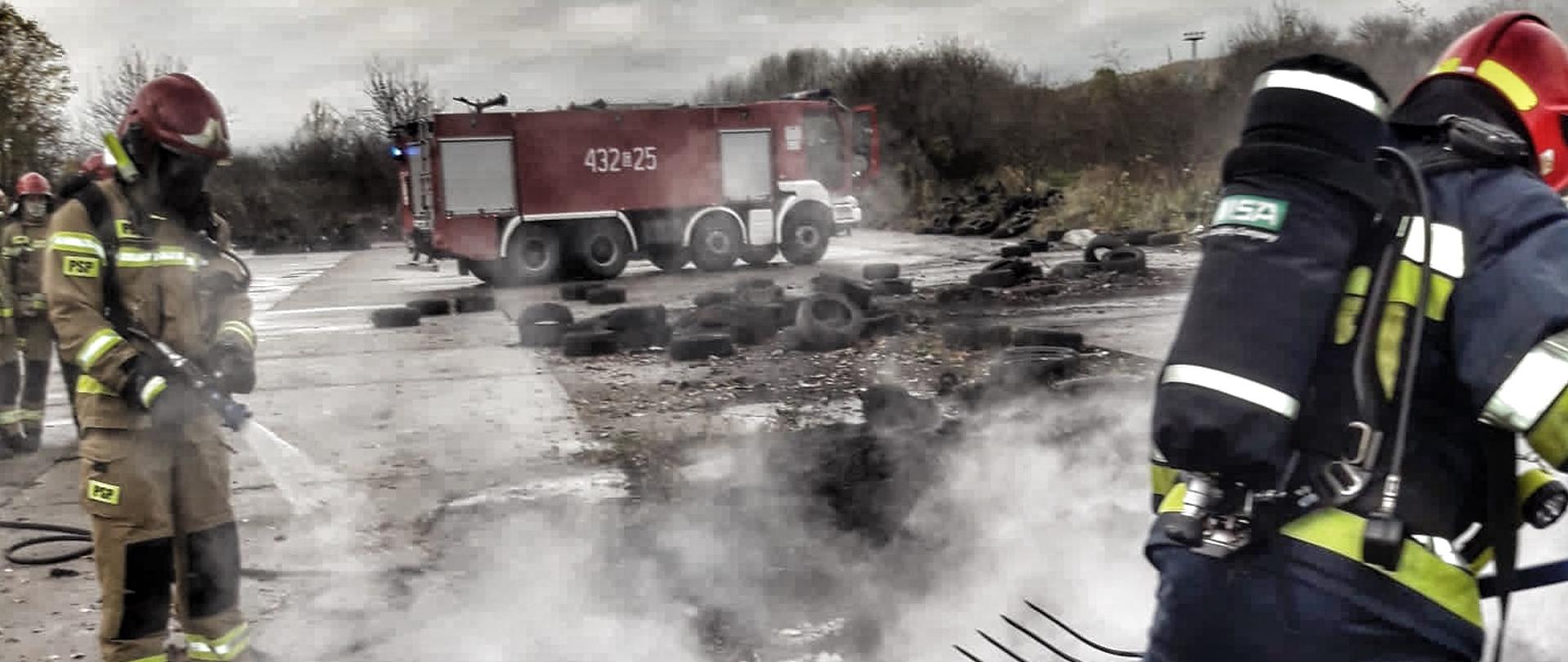 Na betonowym placu dymią się składowane opony. Strażacy dogaszają pożar. W tle stoi samochód pożarniczy.