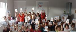 Grafika przedstawia zdjęcie grupowe przedszkolaków w trakcie prowadzenia zajęć profilaktycznych, wszystkie dzieci trzymają w rękach książeczki edukacyjne, pośród nich pracownik OZiPZ