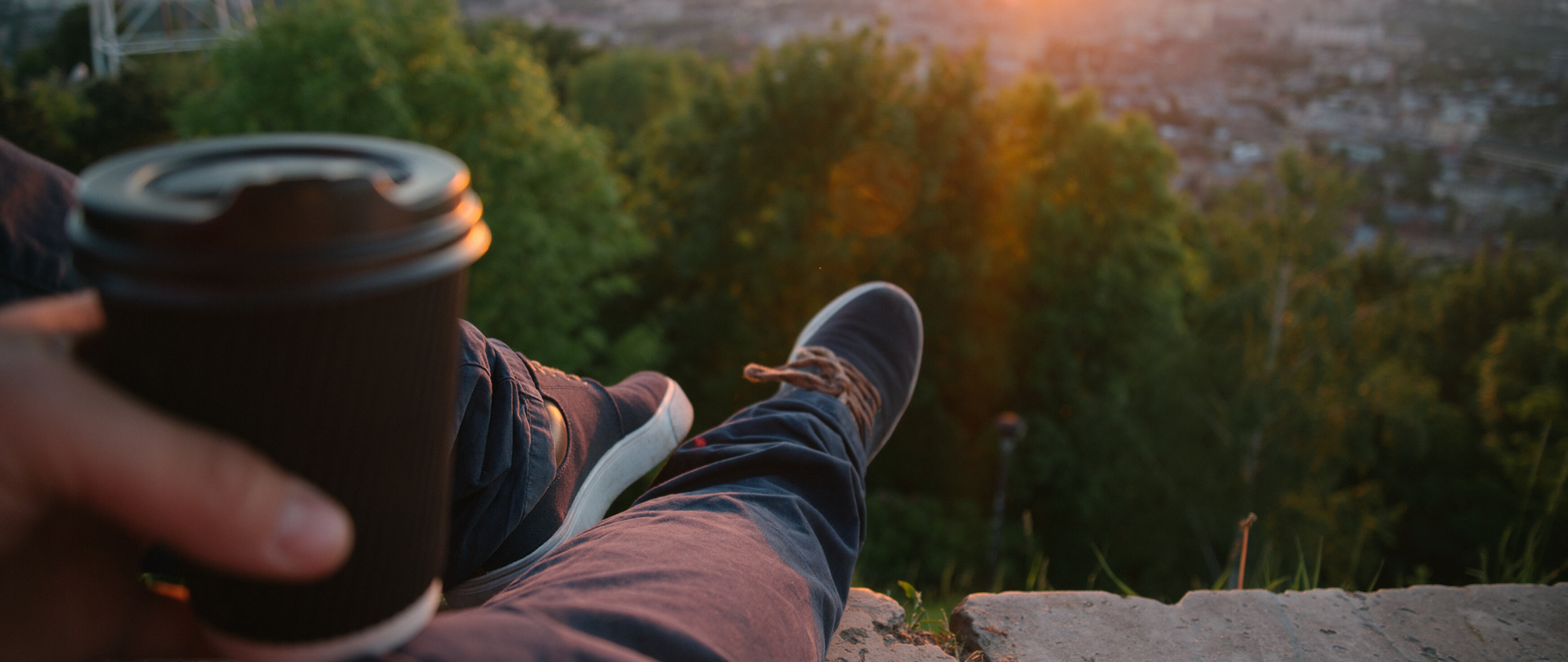 Dłoń mężczyzny trzymająca kubek, widoczne nogi, w tle las oraz góry