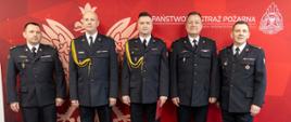 Wspólne zdjęcie oficerów Państwowej Straży Pożarnej na tle czerwonego banneru Państwowej Straży Pożarnej z jasnym Godłem Rzeczypospolitej Polskiej.