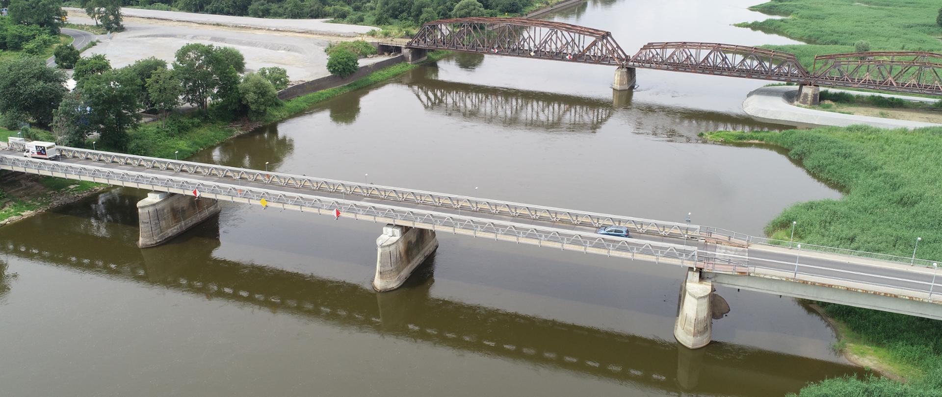 Na zdjęciu widoczne są dwa mosty nad rzeką, jeden drogowy drugi kolejowy, otoczone zielenią.