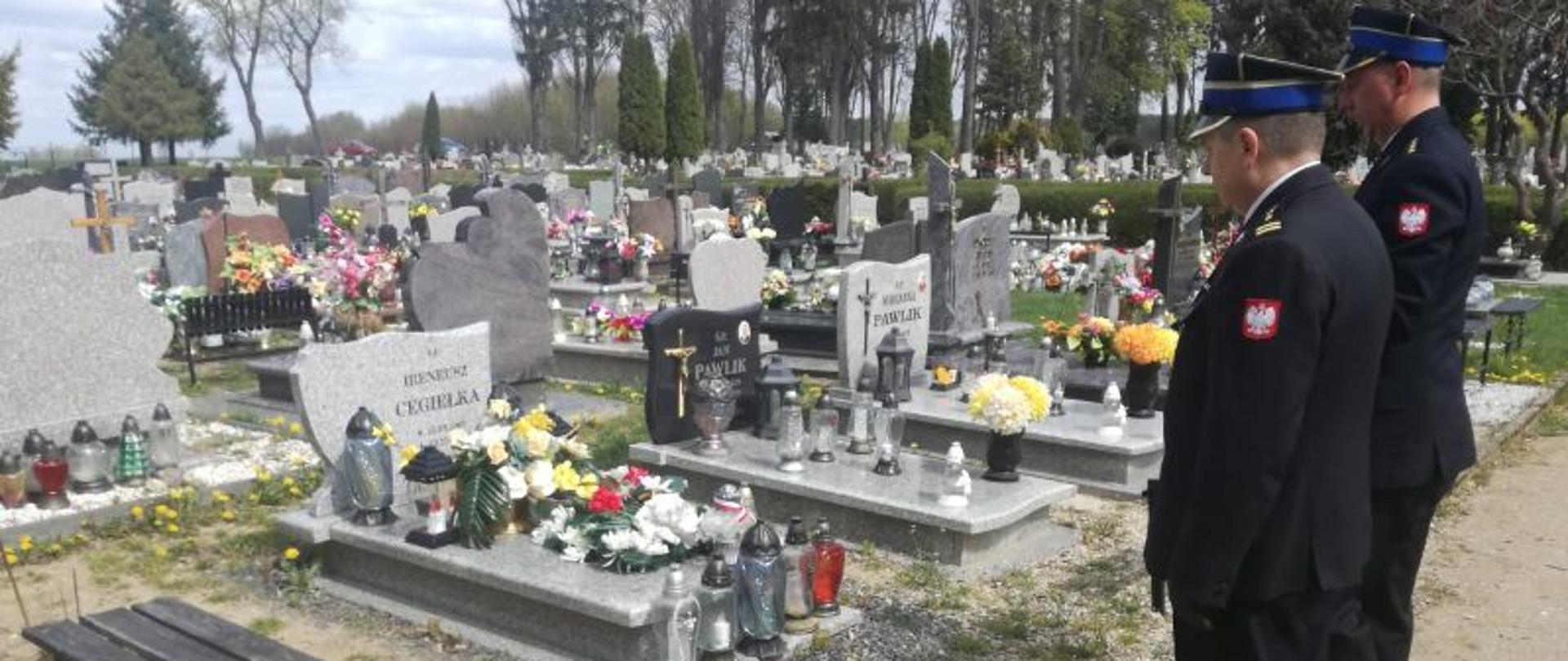 Zdjęcie wykonane w dzień na cmentarzu. Centralny punkt zdjęcia stanowi grób jednego ze strażaków. Przy grobie stoi Komendant i jego zastępca. Oddają hołd zmarłemu strażakowi