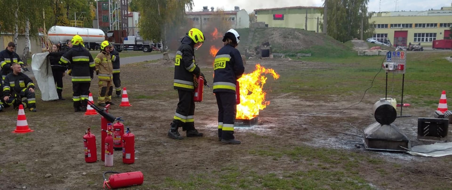 Dwóch strażaków w granatowych ubraniach specjalnych stojących tyłem do osoby robiącej zdjęcie. Obydwaj trzymają w ręku czerwone gaśnice, przed nimi taca metalowa, w której płonie olej napędowy. Strażacy próbują ugasić pożar. Z lewej grupa kilku strażaków również w ubraniach specjalnych przyglądająca się szkoleniu.