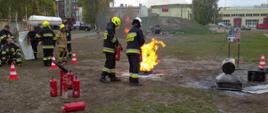 Dwóch strażaków w granatowych ubraniach specjalnych stojących tyłem do osoby robiącej zdjęcie. Obydwaj trzymają w ręku czerwone gaśnice, przed nimi taca metalowa, w której płonie olej napędowy. Strażacy próbują ugasić pożar. Z lewej grupa kilku strażaków również w ubraniach specjalnych przyglądająca się szkoleniu.