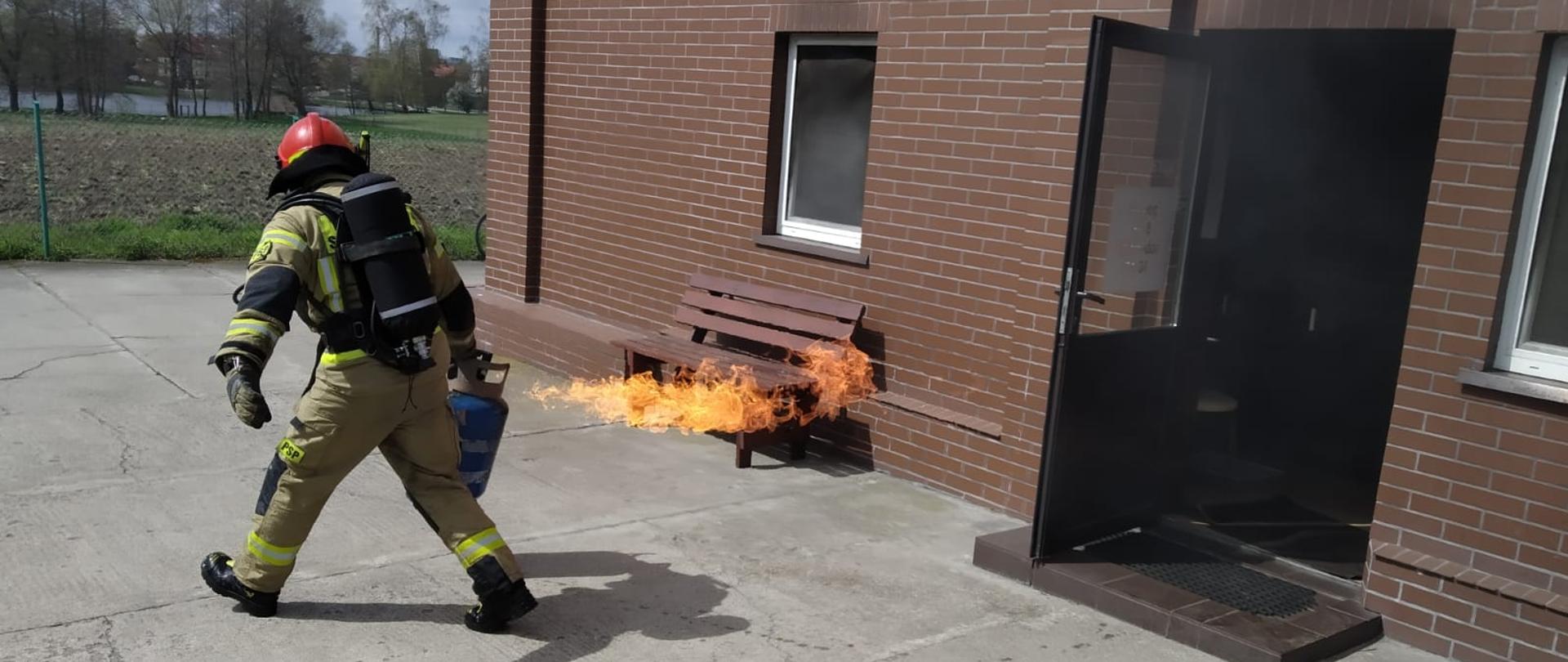 Fotografia przedstawia ujęcie placu betonowego przed budynkiem. Z otwartych drzwi do budynku wydobywa się czarny dym. Przed budynkiem strażak w ubraniu specjalnym, hełmie, z założonym aparatem ochrony dróg oddechowych niesie jedenastokilogramową butlę z gazem propan butan. Butla ma uszkodzony zawór z którego wydobywa się silny, płonący strumień gazu. Jest dzień.