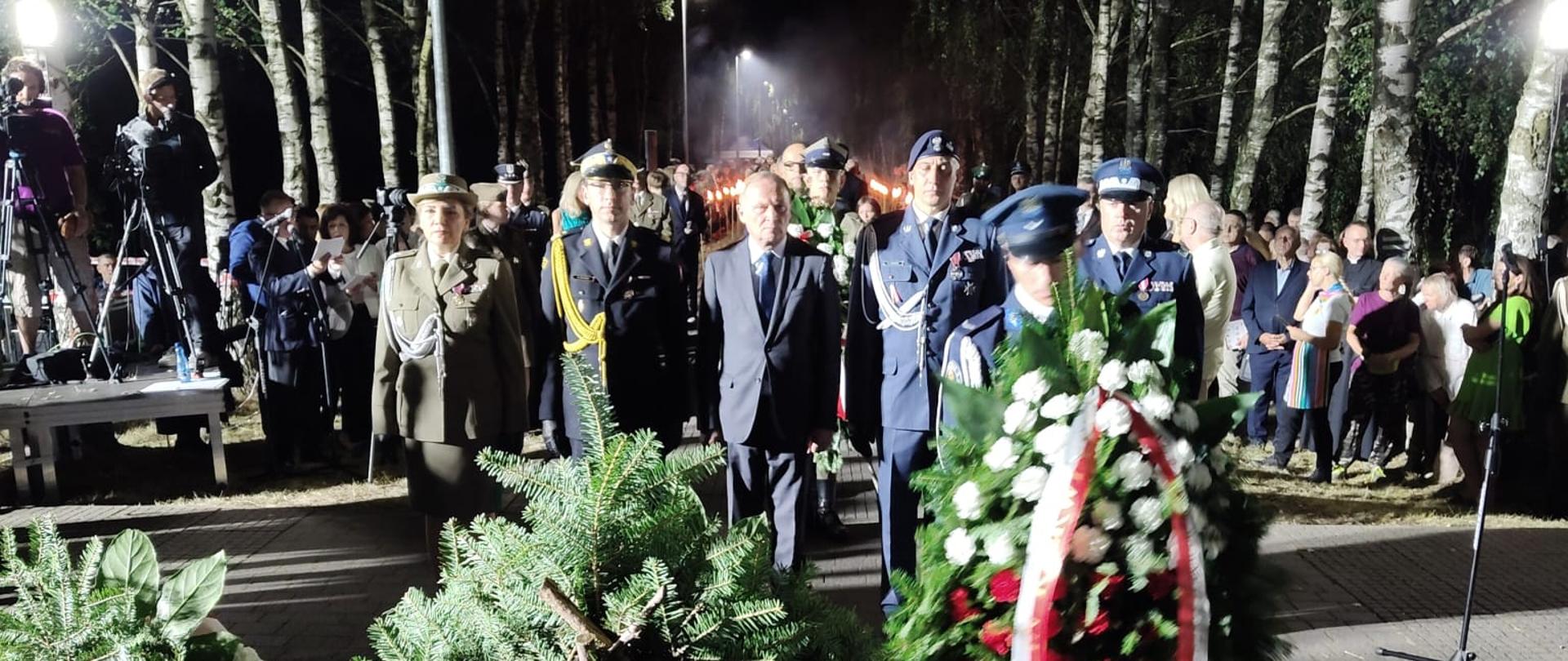 Zdjęcie wykonane w nocy. Przedstawia grupę ludzi, w tym wojskowych w mundurach, idących wzdłuż alei, oświetlonej lampami. Na pierwszym planie znajdują się wieńce i wiązanki kwiatów. Wieńce ozdobione są biało-czerwonymi wstęgami. Wśród uczestników widać osoby w mundurach wojskowych, duchownego oraz cywilów. W tle widać drzewa i tłum.