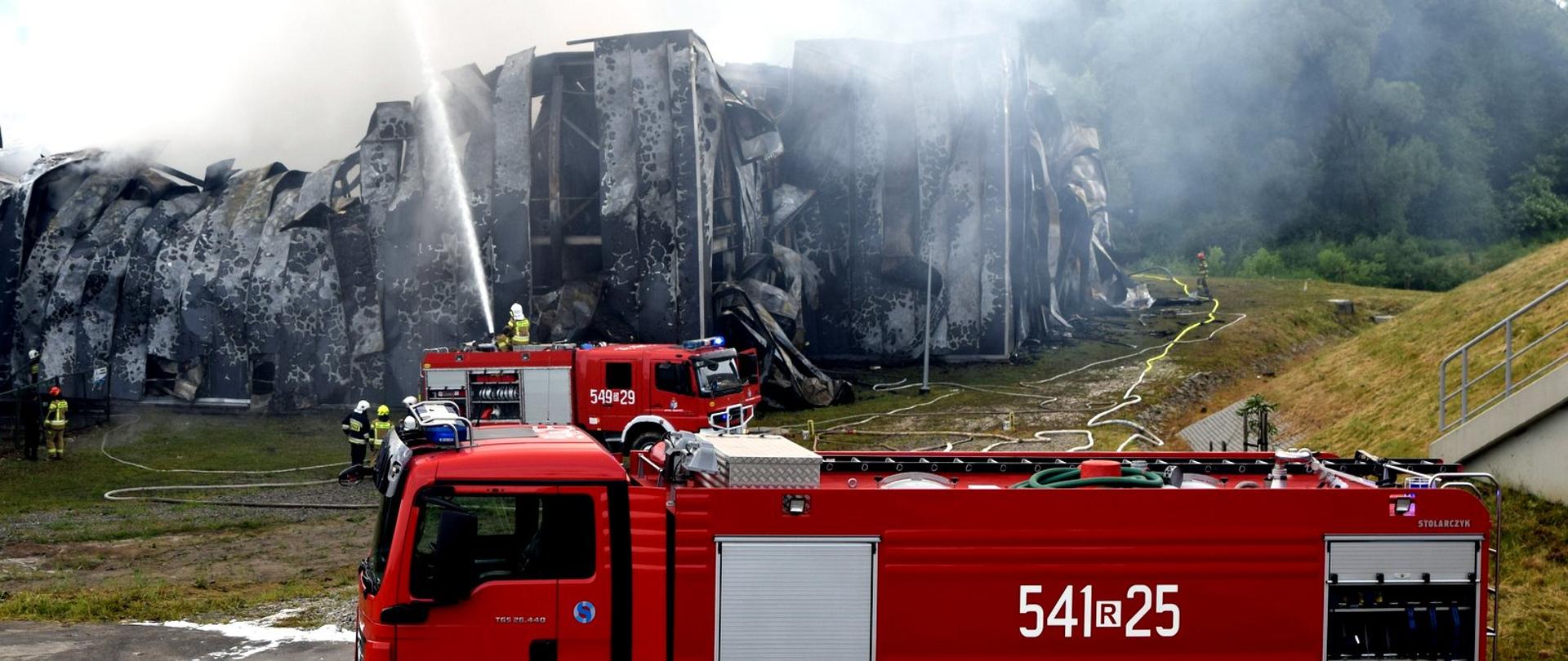 Zdjęcie zrobione na zewnątrz. Na pierwszym planie strażacki samochód gaśniczy. Zanim widać drugi pojazd ratowniczo-gaśniczy strażacy gaszą płonący obiekt.