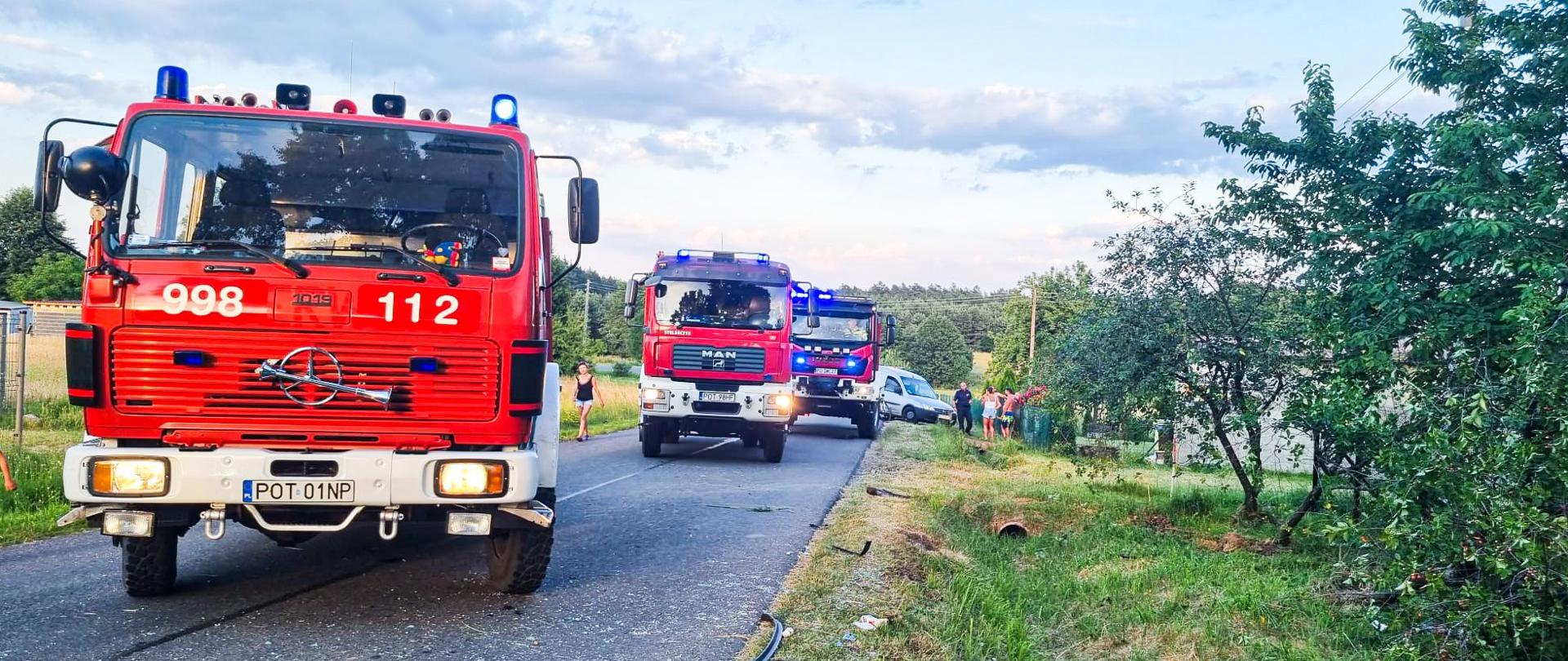 Samochody strażackie stoją na drodze blokując ruch. Na drugim planie widać osoby postronne.