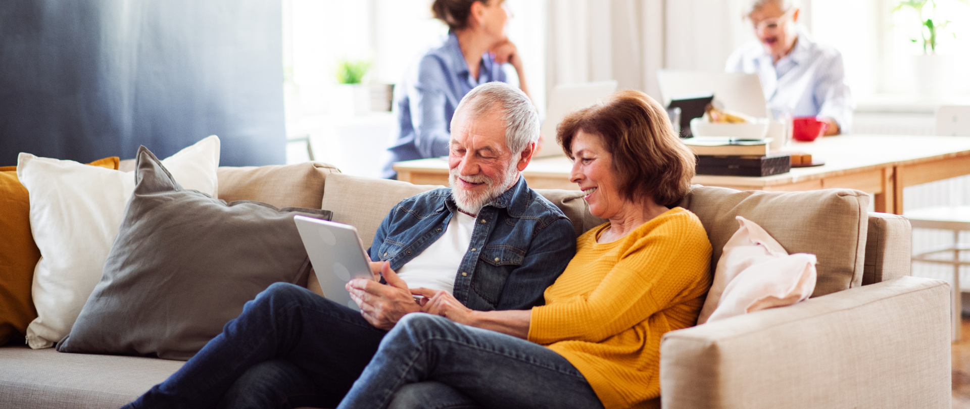 Dwoje seniorów siedzi na kanapie - mężczyzna trzyma w rękach tablet. Kobieta - wskazuje na ekran urządzenia.