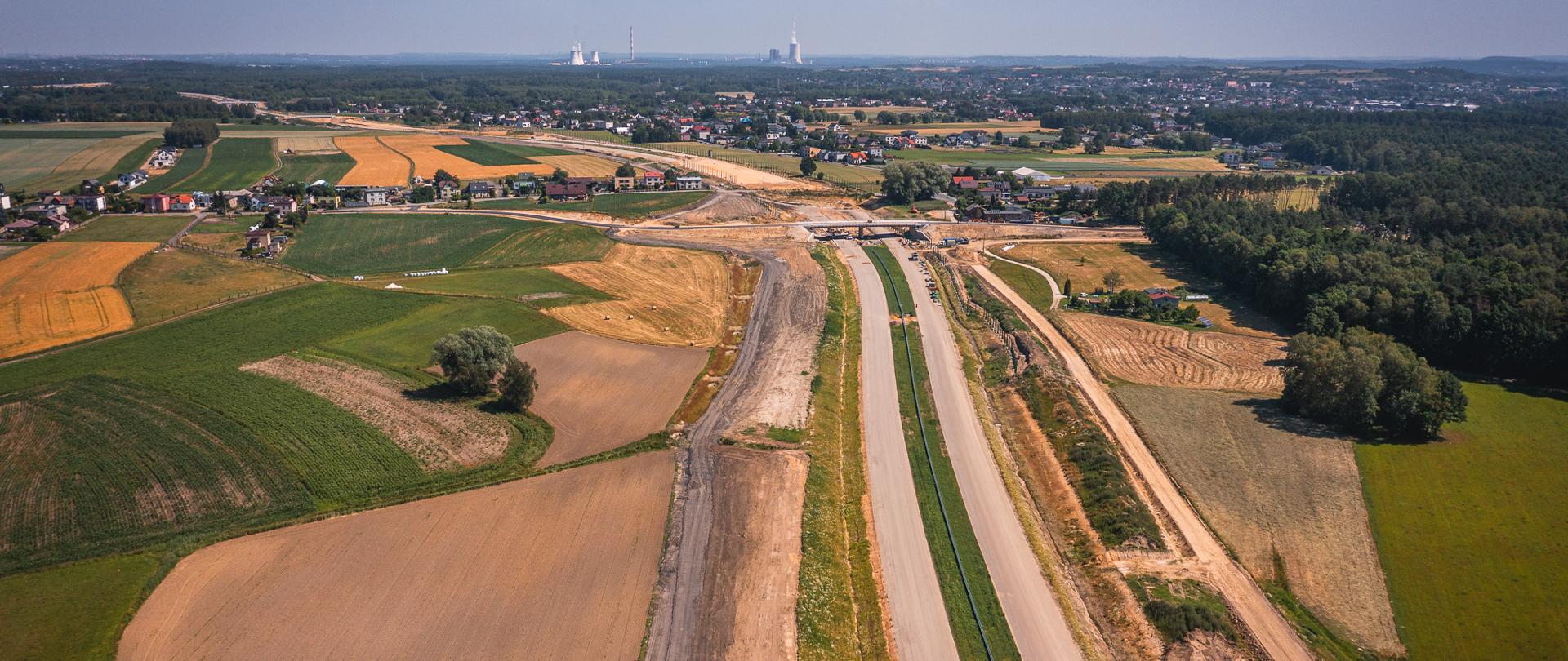 „Widok z lotu ptaka na budowę nowej drogi przebiegającej przez tereny rolnicze. Widoczna jezdnia, nasypy drogowe, wiadukt oraz zabudowa miejscowości w tle.”