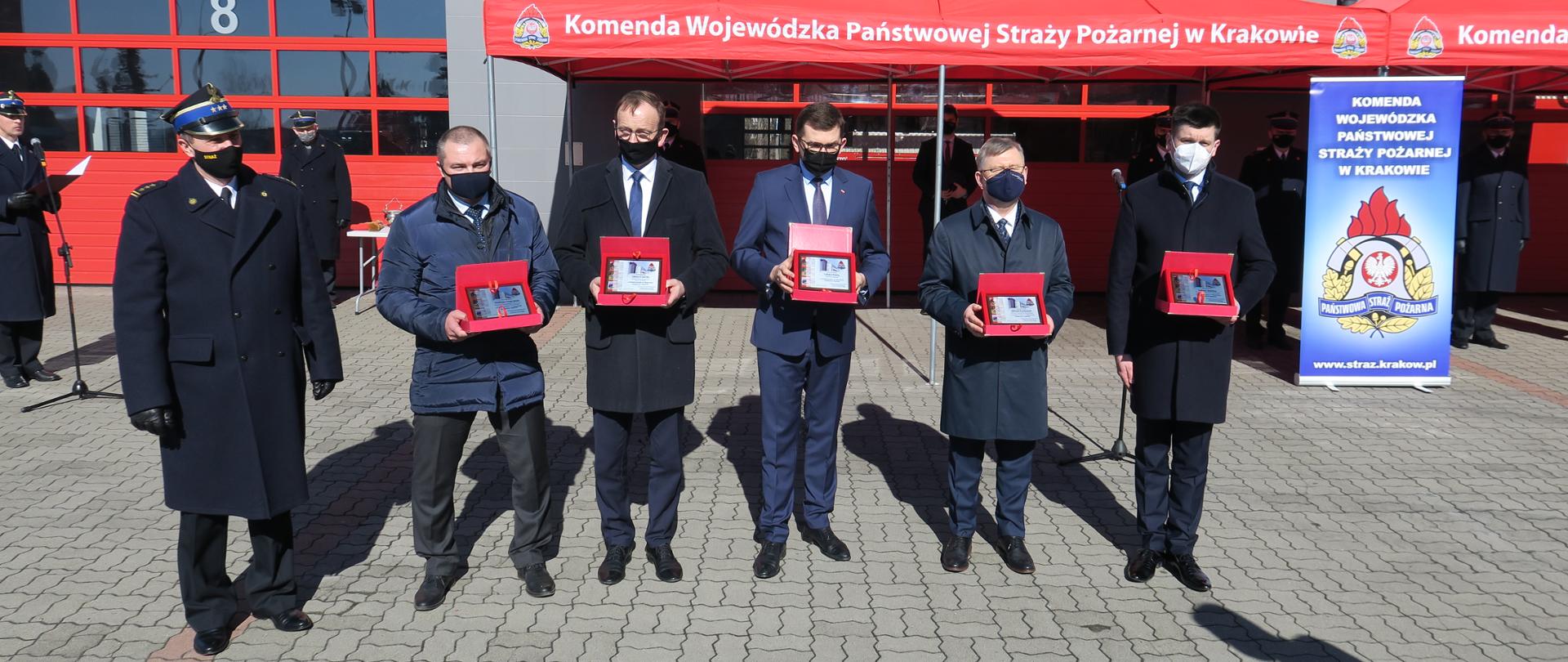 W tle widoczny jest budynek Komendy Wojewódzkiej PSP w Krakowie. Na pierwszym planie od lewej Małopolski Komendant Wojewódzki PSP oraz osoby odbierające okolicznościowe grawertony: dyrektor wydziału z Wojewódzkiego Funduszu Ochrony Środowiska i Gospodarki Wodnej, Wiceminister Klimatu i Środowiska, Wojewoda Małopolski, Marszałek 