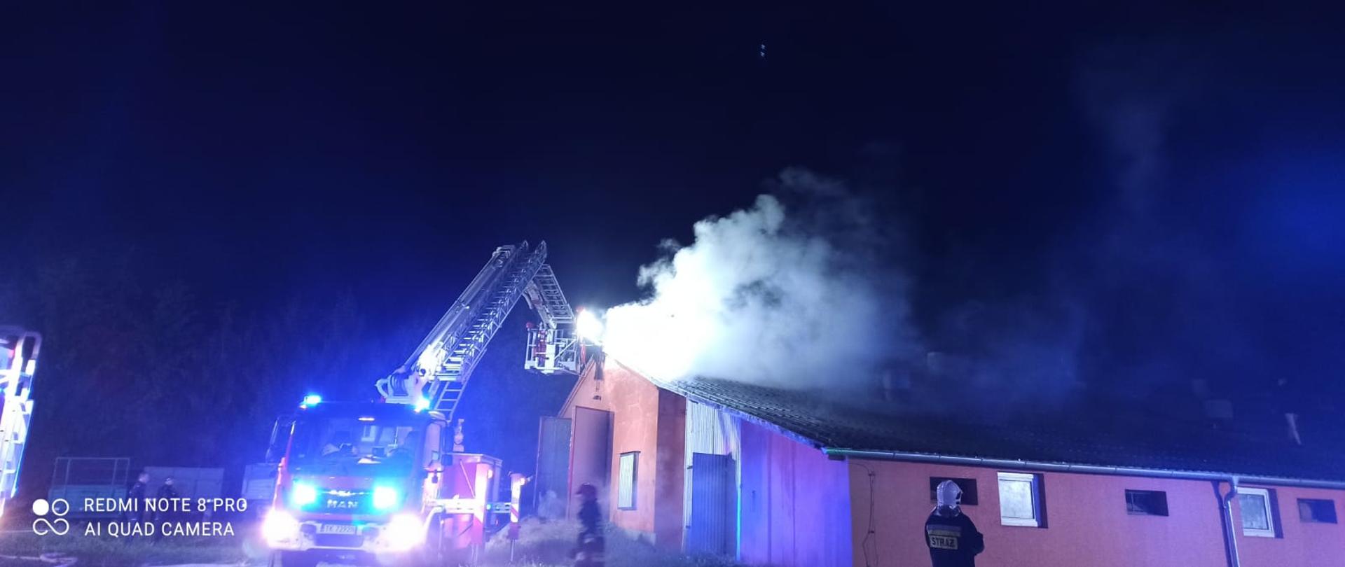 Zdjęcie przedstawia samochód specjalny SHD-23 z rozłożonym podnośnikiem hydraulicznym. W jego koszu znajdują się strażacy zabezpieczeniu w aparaty ochrony dróg oddechowych, którzy gaszą pożar rozprzestrzeniający się na dachu budynku. W miejscu wystąpienia pożaru widoczne także duże zadymienie. Przed budynkiem znajduje się kolejny strażak w białym hełmie oraz umundurowaniu specjalnym. 