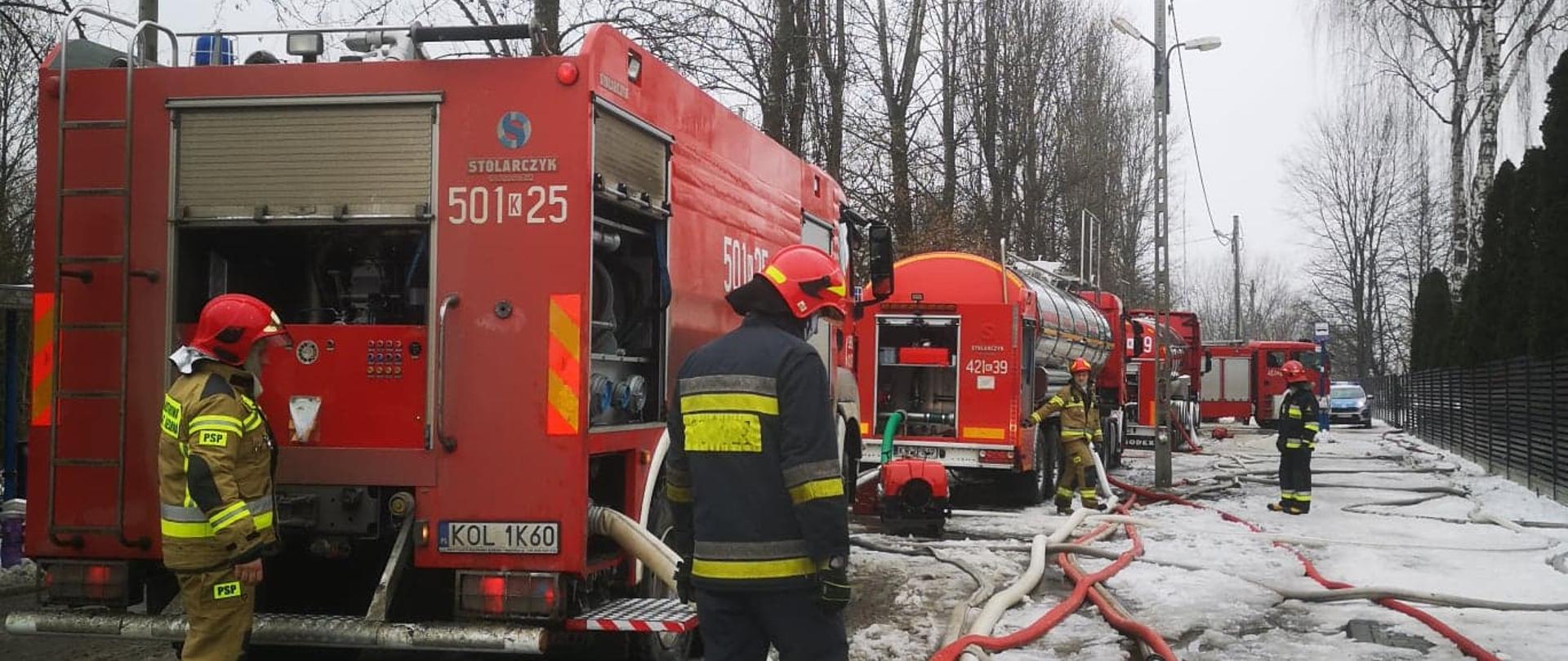 Strażacy w ubraniach specjalnych wśród węży pożarniczych w tle samochody gaśnicze oraz cysterna dostarczające wodę