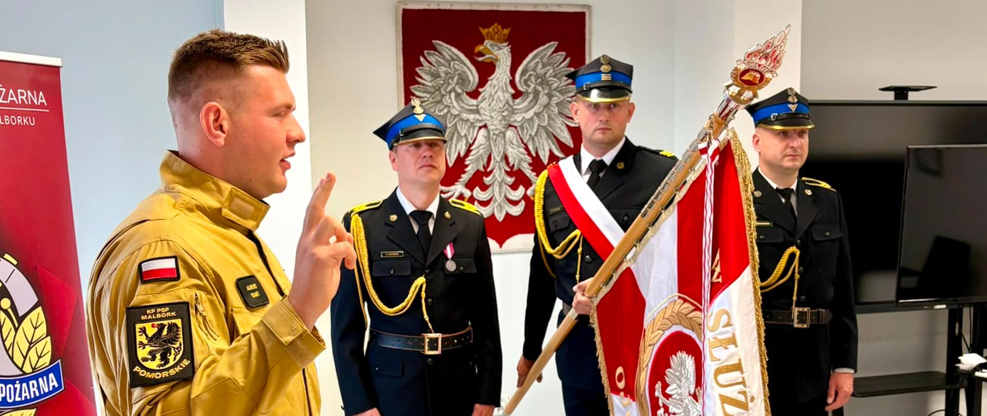 Na zdjęciu widoczna jest uroczystość odbywająca się w pomieszczeniu służbowym Państwowej Straży Pożarnej.
Na pierwszym planie, po lewej stronie, stoi strażak ubrany w piaskowy ubiór specjalny. Ma uniesioną prawą rękę z dwoma wyprostowanymi palcami, co wskazuje, że najprawdopodobniej składa rotę ślubowania lub przyrzeczenia. Jego sylwetka jest zwrócona w stronę pocztu sztandarowego.
Na drugim planie znajduje się trzyosobowy poczet sztandarowy w mundurach wyjściowych. Środkowy strażak trzyma sztandar jednostki, natomiast dwaj pozostali pełnią funkcję asysty. Wszyscy zachowują postawę zasadniczą.
Na ścianie za pocztem sztandarowym znajduje się godło Rzeczypospolitej Polskiej w czerwonym tle. Po lewej stronie kadru widoczny jest fragment roll-upu z logo Państwowej Straży Pożarnej i napisem odnoszącym się do Komendy Powiatowej PSP w Malborku. Po prawej stronie znajduje się wyłączony telewizor ustawiony na stojaku.
Pomieszczenie jest jasne, z jasną podłogą i oświetleniem sufitowym, co nadaje fotografii oficjalny i uporządkowany charakter.