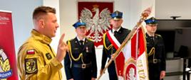 Na zdjęciu widoczna jest uroczystość odbywająca się w pomieszczeniu służbowym Państwowej Straży Pożarnej.
Na pierwszym planie, po lewej stronie, stoi strażak ubrany w piaskowy ubiór specjalny. Ma uniesioną prawą rękę z dwoma wyprostowanymi palcami, co wskazuje, że najprawdopodobniej składa rotę ślubowania lub przyrzeczenia. Jego sylwetka jest zwrócona w stronę pocztu sztandarowego.
Na drugim planie znajduje się trzyosobowy poczet sztandarowy w mundurach wyjściowych. Środkowy strażak trzyma sztandar jednostki, natomiast dwaj pozostali pełnią funkcję asysty. Wszyscy zachowują postawę zasadniczą.
Na ścianie za pocztem sztandarowym znajduje się godło Rzeczypospolitej Polskiej w czerwonym tle. Po lewej stronie kadru widoczny jest fragment roll-upu z logo Państwowej Straży Pożarnej i napisem odnoszącym się do Komendy Powiatowej PSP w Malborku. Po prawej stronie znajduje się wyłączony telewizor ustawiony na stojaku.
Pomieszczenie jest jasne, z jasną podłogą i oświetleniem sufitowym, co nadaje fotografii oficjalny i uporządkowany charakter.