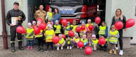 Na zdjęciu widać grupę przedszkolaków z opiekunami i strażakami, dzieci trzymają balony, w tle samochód pożarniczy
