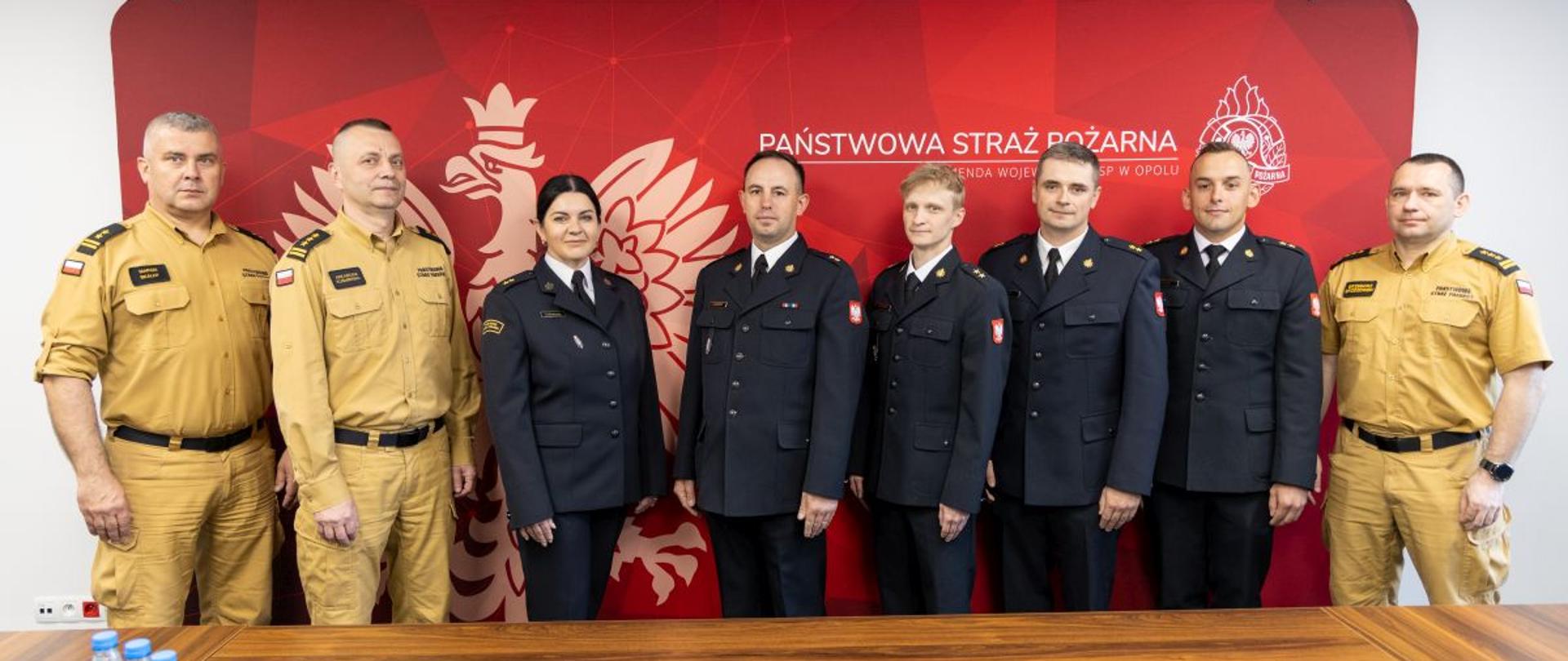 Ośmiu funkcjonariuszy stoi w rzędzie na tle czerwonej ścianki z białym orłem i napisem „Państwowa Straż Pożarna – Komenda Wojewódzka PSP w Opolu”. Pięć osób ma granatowe mundury galowe, trzy osoby jasne mundury służbowe. Przed nimi stoi stół z butelkami wody, pilotem i klawiaturą. Zdjęcie dokumentuje wspólne spotkanie kierownictwa PSP z nowo promowanymi młodszymi kapitanami.