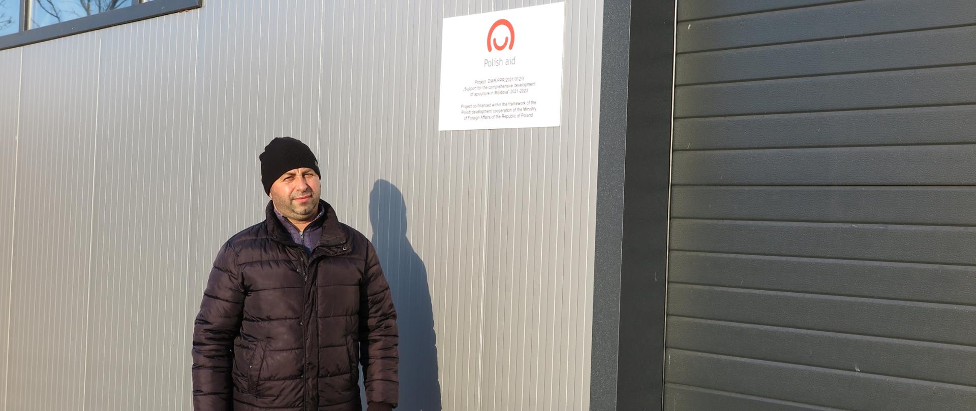 Mężczyzna w czapce pozuje na tle szarej ściany. Po jego lewej stronie na ścianie znajduje się czerwone logo polskiej pomocy na białym tle.