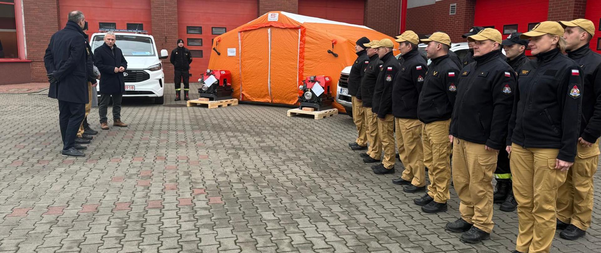 Zdjęcie przedstawia Strażaków z Komendy Miejskiej Państwowej Straży Pożarnej w Piekarach Śląskich, Posła na Sejm Rzeczypospolitej Polskiej Łukasza Ściebiorowskiego, Zastępcę Prezydenta Miasta Piekary Śląskie Krzysztofa Ślusarczyka, Wiceprzewodniczącą Rady Miasta Piekary Śląskie Klaudie Franiel, Komendanta Miejskiego Państwowej Straży Pożarnej w Piekarach Śląskich starszego brygadiera Tomasza Dziechciarza, namiot pneumatyczny, samochód kwatermistrzowski, samochód rozpoznawczo-ratowniczy, motopompy do wody czystej.