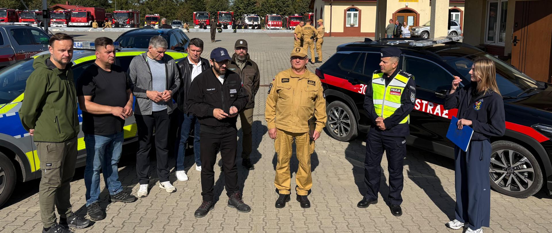 Przedstawiciele Państwowej Straży Pożarnej, Policji, innych służb i instytucji uczestniczą w briefingu prasowym podczas ćwiczeń powiatowych. Na pierwszym planie stoi grupa osób, w tym komendant miejski PSP w Rzeszowie w piaskowym umundurowaniu służbowym, funkcjonariusz Policji w kamizelce odblaskowej oraz kobieta trzymająca niebieską teczkę. Po obu stronach grupy zaparkowane są samochody służbowe Policji i Państwowej Straży Pożarnej. W głębi placu ustawiono liczne samochody ratowniczo-gaśnicze.
