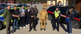 Przedstawiciele Państwowej Straży Pożarnej, Policji, innych służb i instytucji uczestniczą w briefingu prasowym podczas ćwiczeń powiatowych. Na pierwszym planie stoi grupa osób, w tym komendant miejski PSP w Rzeszowie w piaskowym umundurowaniu służbowym, funkcjonariusz Policji w kamizelce odblaskowej oraz kobieta trzymająca niebieską teczkę. Po obu stronach grupy zaparkowane są samochody służbowe Policji i Państwowej Straży Pożarnej. W głębi placu ustawiono liczne samochody ratowniczo-gaśnicze.