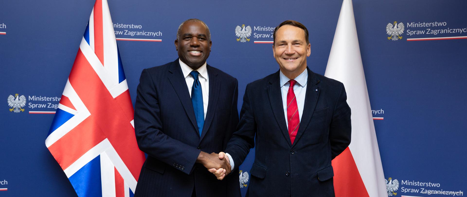 Na zdjęciu jest dwóch mężczyzn w ciemnych garniturach. Podają sobie dłonie. Patrzą w obiektyw i uśmiechają się. Za nimi są: niebieska ścianka z logo MSZ oraz dwie flagi - Wielkiej Brytanii i Polski.