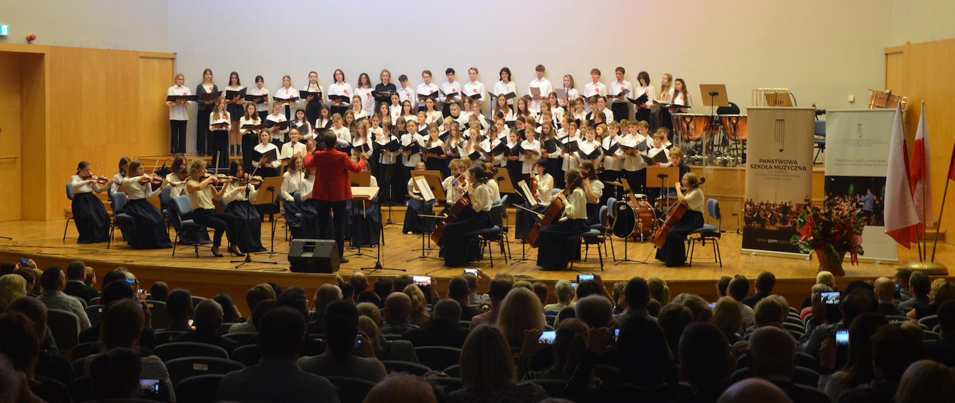 Zdjęcie przedstawia występ orkiestry szkolnej i chóru na scenie Filharmonii Zielonogórskiej podczas Koncertu z okazji Narodowego Święta Niepodległości