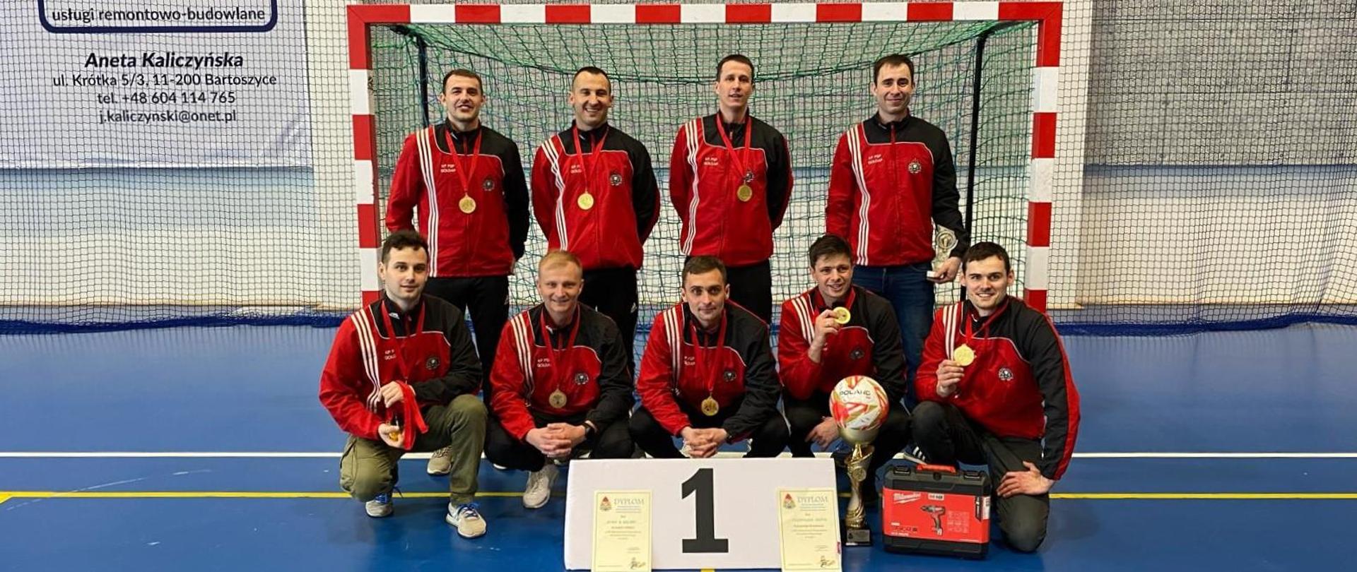 Zdjęcie przedstawia drużynę Komendy Powiatowej Państwowej Straży Pożarnej w Gołdapi (dziewięciu zawodników) - Mistrzów Województwa Warmińsko-Mazurskiego w Futsalu ze zdobytymi trofeami, na tle bramki w hali widowiskowo-sportowej Bartoszyckiego Ośrodka Sportu i Rekreacji