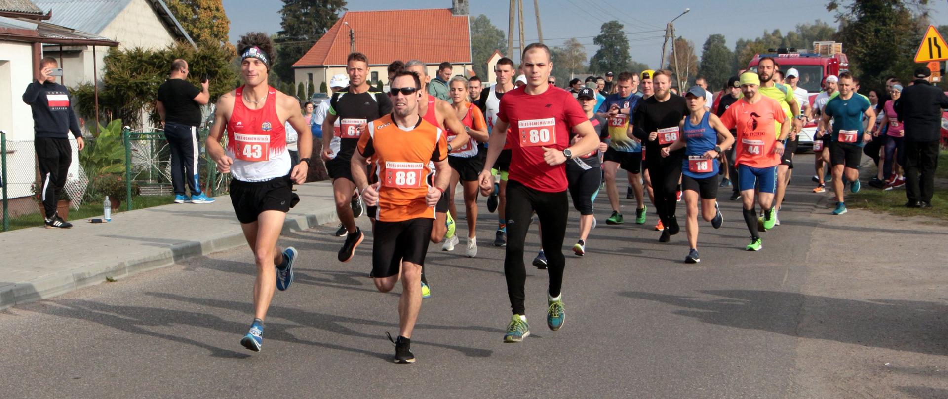 Zdjęcie przedstawia drogę asfaltową, którą biegną osoby ubrane w wielokolorowe ciuchy. Każdy ma przypięty numer identyfikacyjny. W tle domy zabudowy jednorodzinnej.