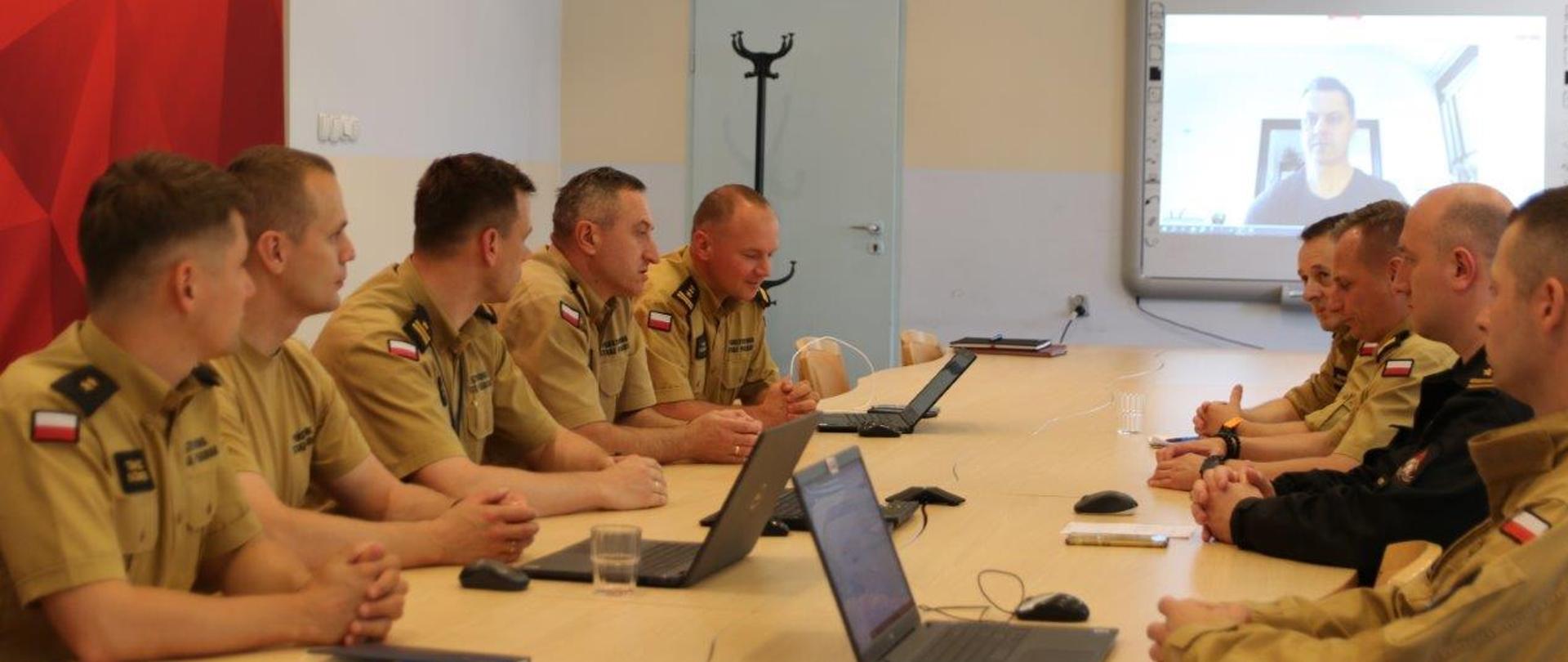 Członkowie zespołu siedzą przy stole w sali , na monitorze telewizora widoczny wizerunek uczestnika w trybie wideokonferencji